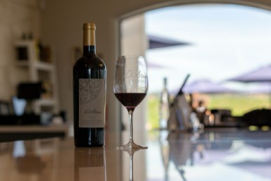 Dark wine bottle and wine glass with red wine sitting on a white countertop reflecting the sunshine from outside a window